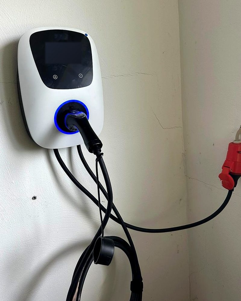 Wei&szlig;e Wallbox mit Ladekabel an einer Garagenwand, rechts ein roter Drehstromstecker