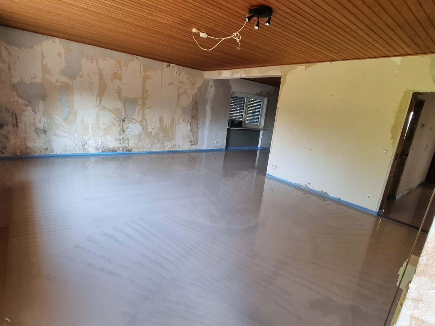Frischer Flie&szlig;estrich in einem Altbau-Wohnraum: Holzdecke mit Lampenkabel, W&auml;nde mit abgel&ouml;sten Tapetenresten, am Wandfu&szlig; der blaue Randd&auml;mmstreifen
