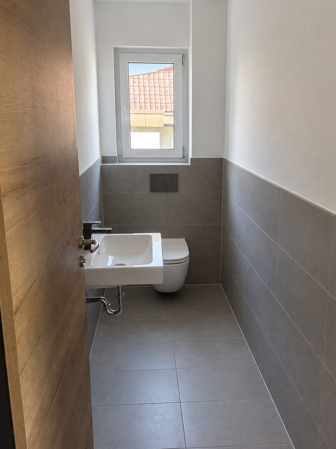 Dasselbe G&auml;ste-WC fertig gefliest: graue Gro&szlig;formatfliesen, Waschbecken, H&auml;nge-WC und Fenster
