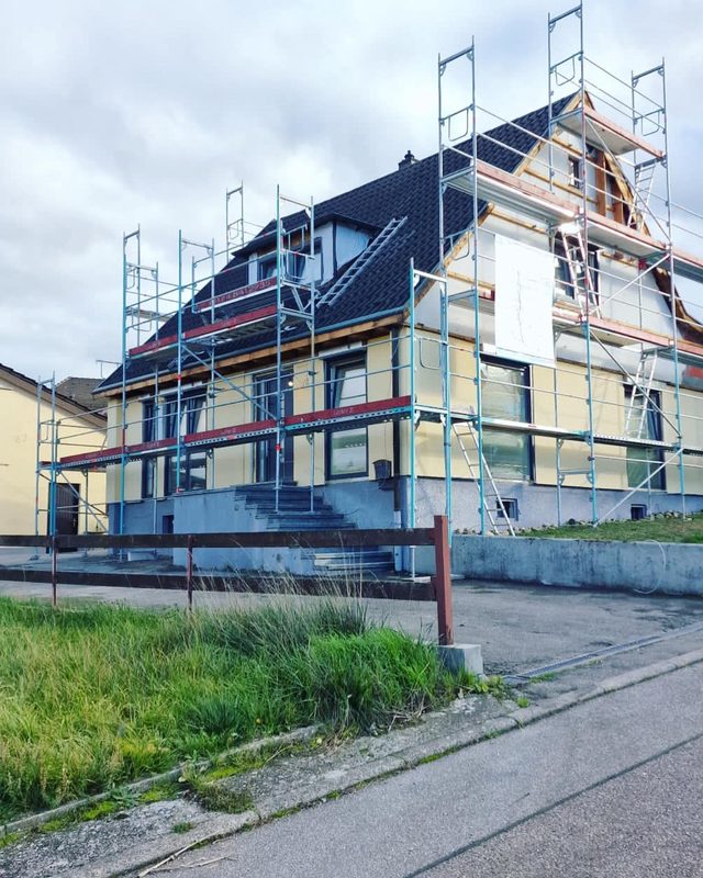 Einfamilienhaus mit dunklem Dach, an Fassade und Giebel einger&uuml;stet, davor Treppe und Wiese