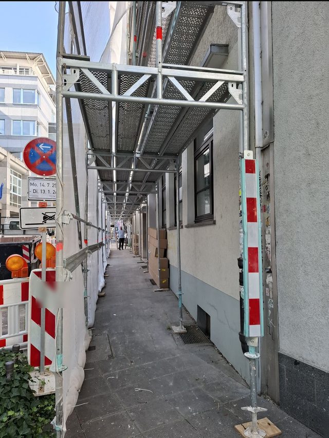 Gehweg unter einem Ger&uuml;st, dar&uuml;ber ein Schutzdach, vorn rot-wei&szlig;e Absperrung und ein Verkehrsschild