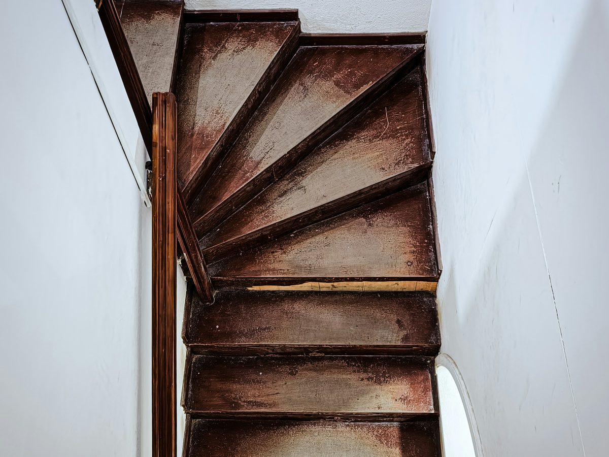 Alte gewendelte Holztreppe mit abgetretenem, rotbraunem Anstrich zwischen wei&szlig;en W&auml;nden
