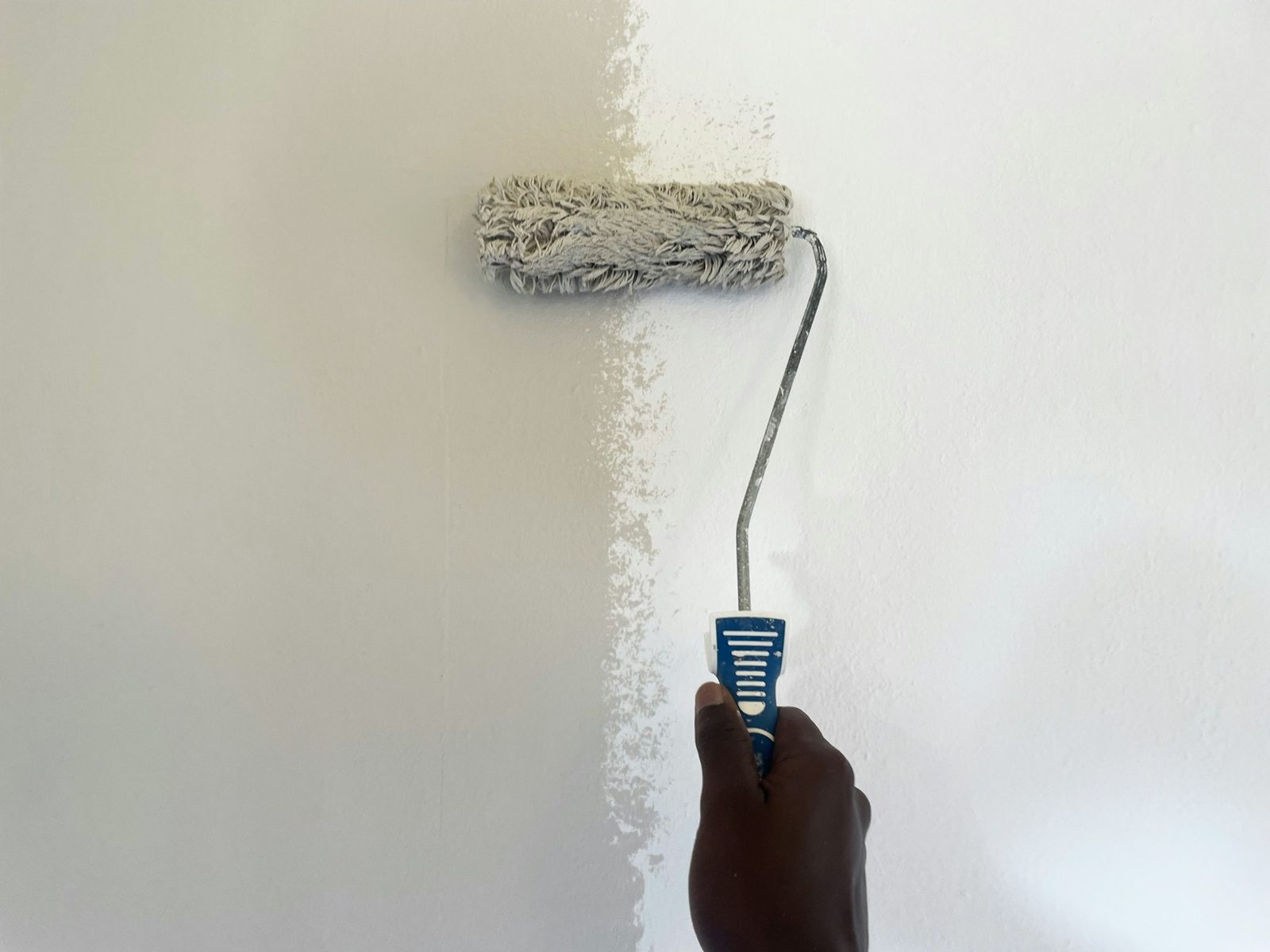 Eine Hand führt eine Farbrolle über eine Wand, links der alte beige Anstrich, rechts das frische Weiß